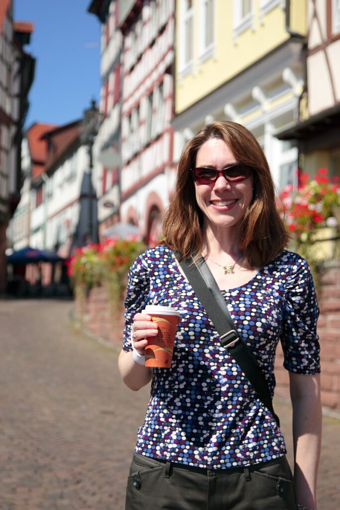 A woman in a patterned shirt and sunglasses enjoys a drink while walking through a picturesque street lined with traditional European architecture.