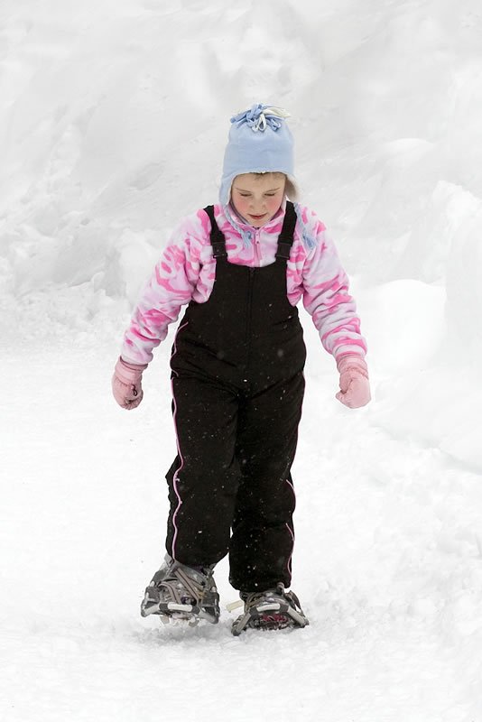 A child wearing a blue hat, pink jacket, and black overalls walks through deep snow using snowshoes.