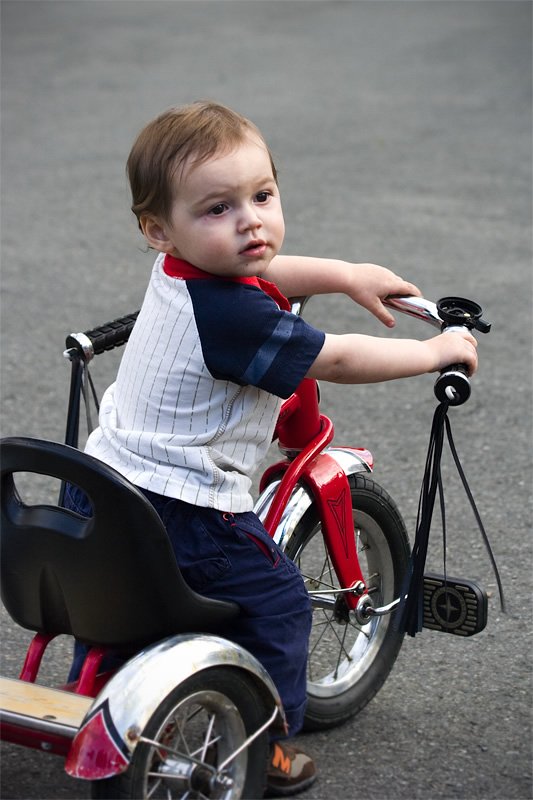 A young boy with light brown hair sits on a red tricycle, gripping the handlebars and looking ahead. He wears a striped shirt and dark pants, preparing to pedal along a paved surface.