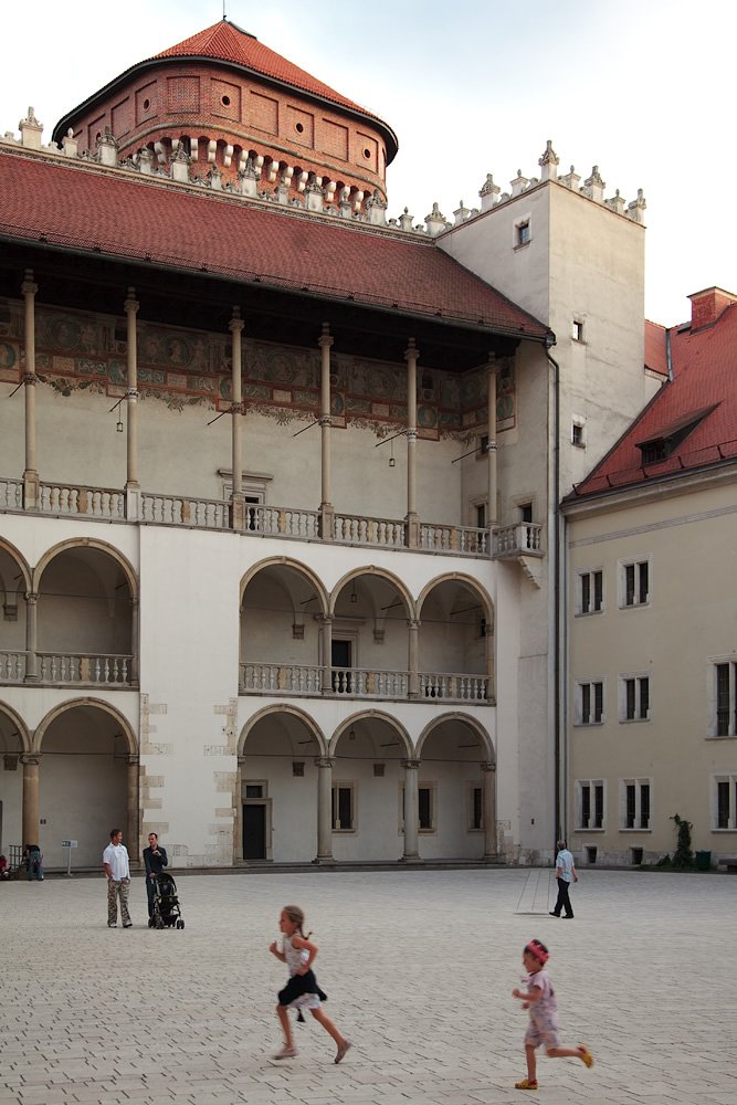 Two children run joyfully across a sunlit stone courtyard surrounded by elegant Renaissance-style buildings with arched walkways and terracotta roofs.