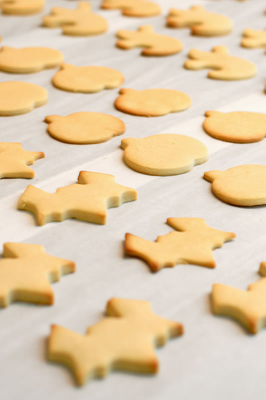 Sugar cookies in various shapes like bears, fish, and Santa hats are arranged on a parchment-lined baking sheet. The cookies are golden brown and ready to be decorated.