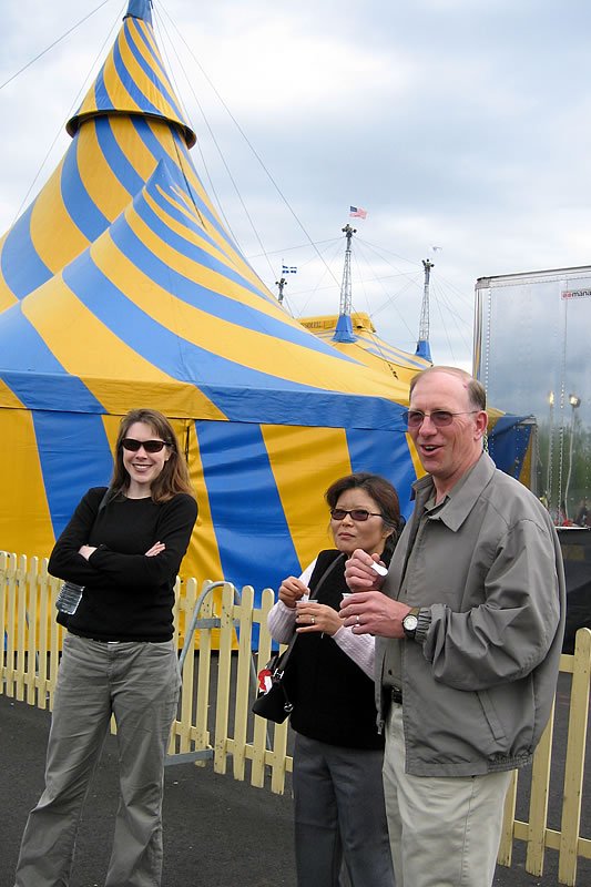 Three individuals standing in front of a vibrant blue and yellow striped circus tent. One person is smiling with arms crossed.