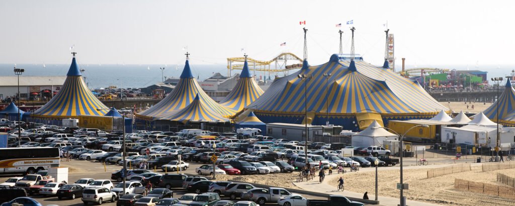 A panoramic view of colorful circus tents set up near the ocean with a fairground in the background.