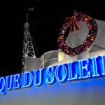 Animal Kingdom A brightly lit blue neon sign for Cirque du Soleil stands against a dark night sky with a festive wreath hanging above it.