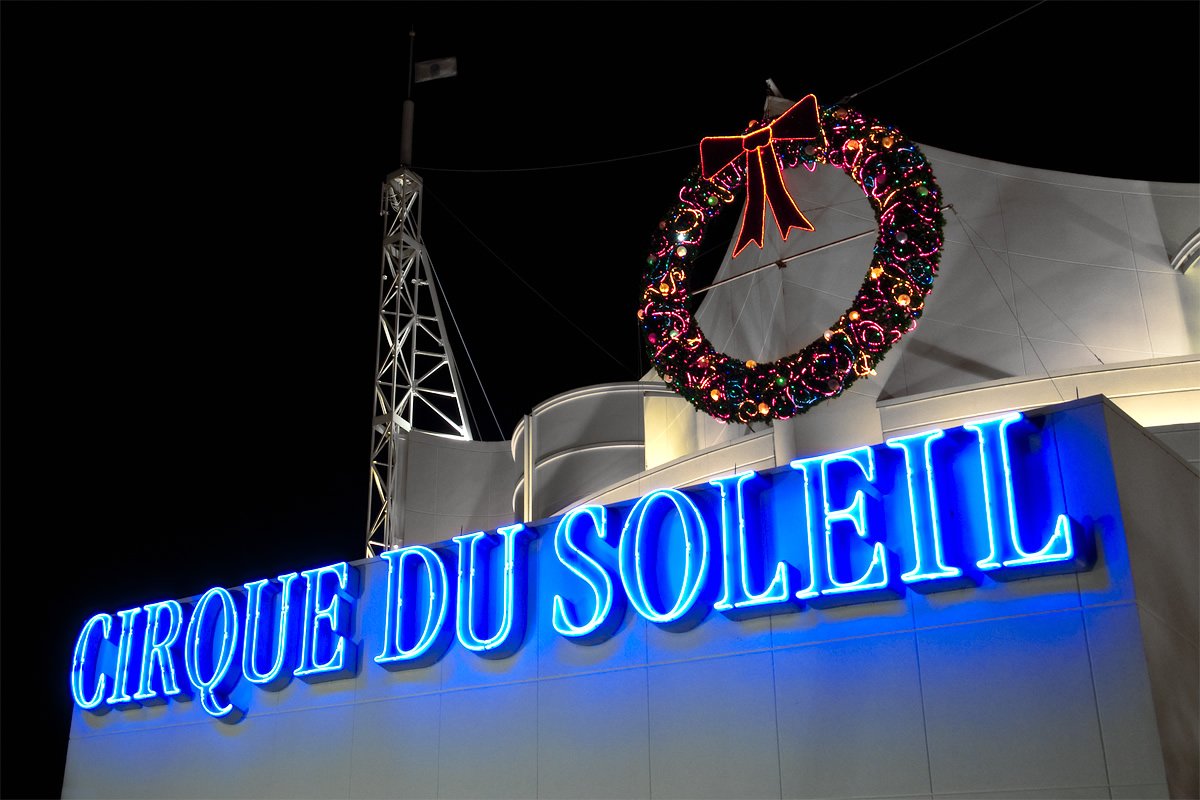 A brightly lit blue neon sign for Cirque du Soleil stands against a dark night sky with a festive wreath hanging above it.