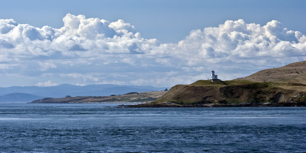 A serene coastal scene featuring a lighthouse perched on a grassy headland under expansive blue skies with fluffy clouds.