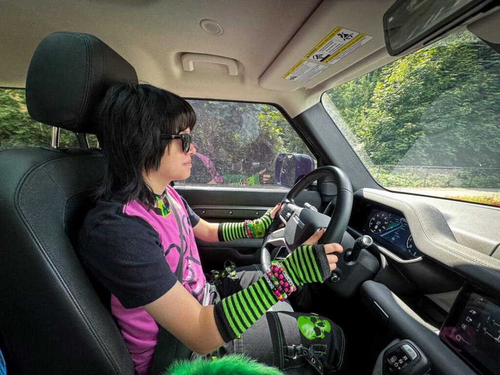 A person wearing a pink and black outfit with green and black striped fingerless gloves is driving a car. The interior of the car is visible, with green trees outside the window.
