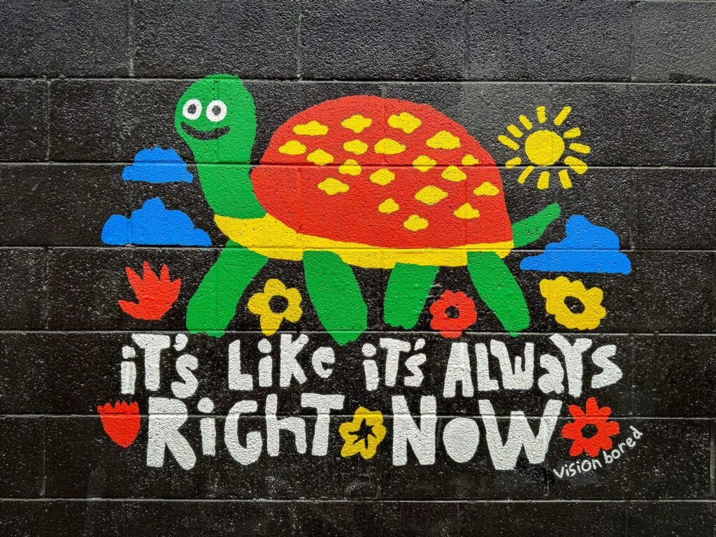 A vibrant mural featuring a green turtle with a red shell filled with yellow spots, surrounded by colorful flowers and clouds under a bright sun. The text reads 'It's Like It's Always Right Now' in playful lettering.