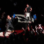 A male singer passionately performing on stage with a drummer in the background, surrounded by an enthusiastic crowd raising their hands.