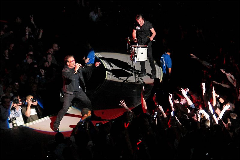 A male singer passionately performing on stage with a drummer in the background, surrounded by an enthusiastic crowd raising their hands.