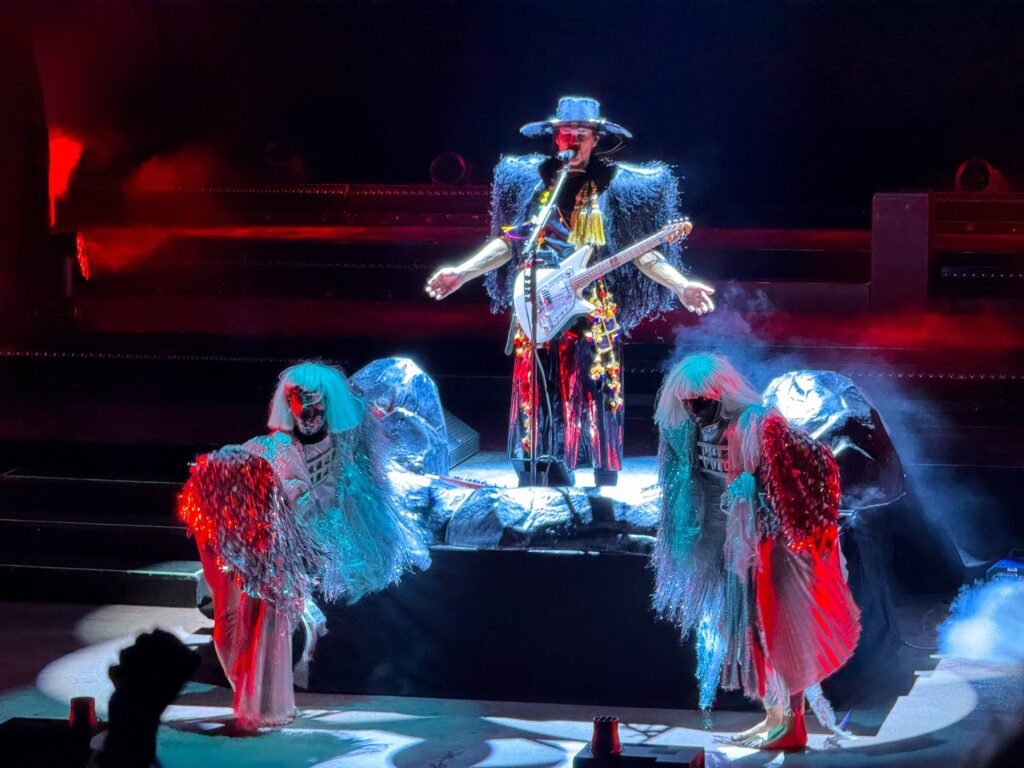 A musician plays guitar on stage surrounded by performers in elaborate costumes.
