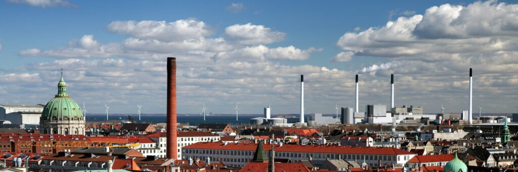 A panoramic view of Copenhagen showcasing golden domes, red-tiled roofs, and prominent chimneys under a partly cloudy sky. The cityscape blends historic architecture with modern elements near the harbor.