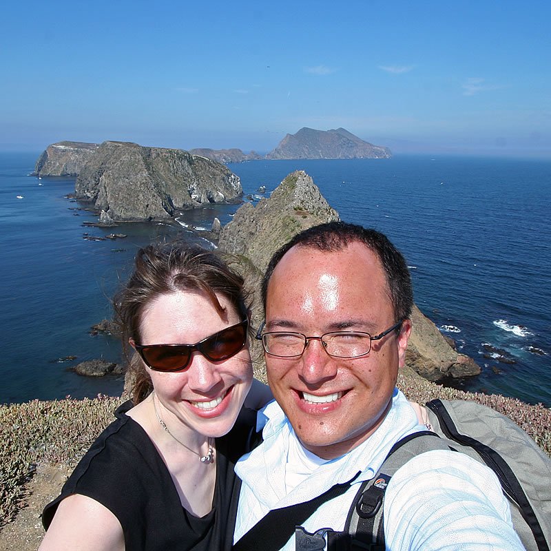 A couple, John and Kristie, smile for a selfie with a stunning coastal landscape in the background.