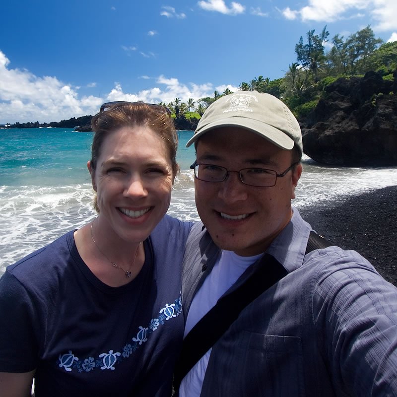 A couple smiles for a selfie on a sunny day at the beach. The man wears a cap and glasses while the woman has her hair tied back.
