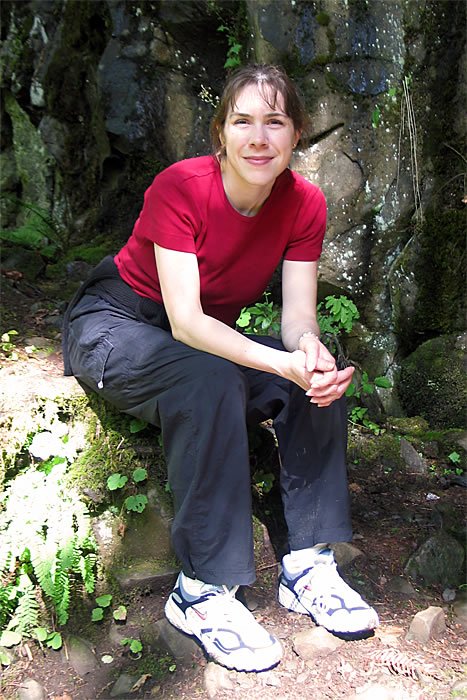 A couple sits on a rocky ledge in a forest. The person on the left wears a red shirt and black pants, smiling at the camera.