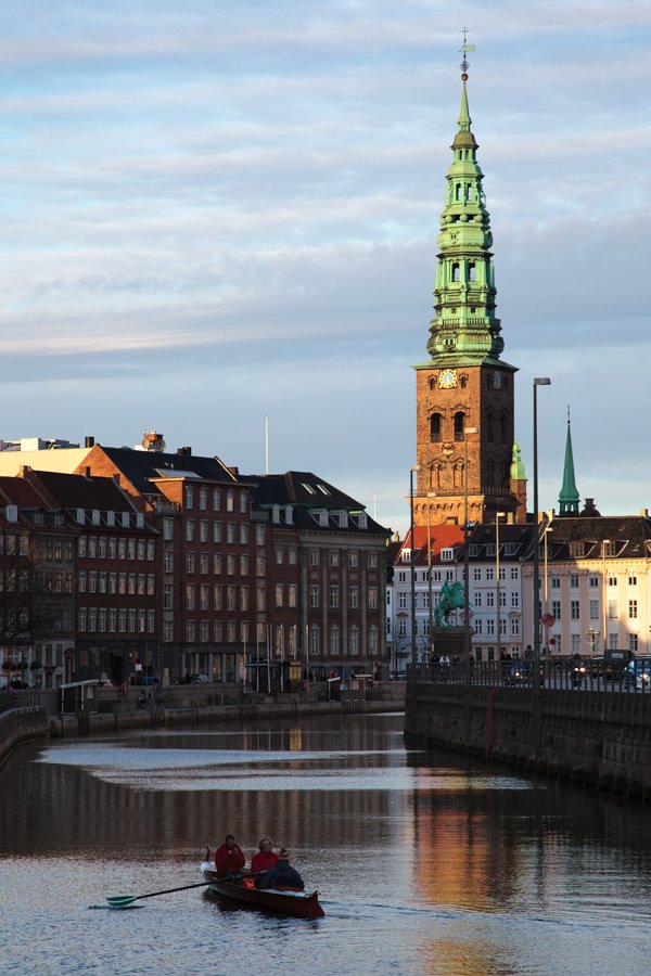 Two people rowing a small boat on a canal, with the iconic green-spired Copenhagen Town Hall in the background.