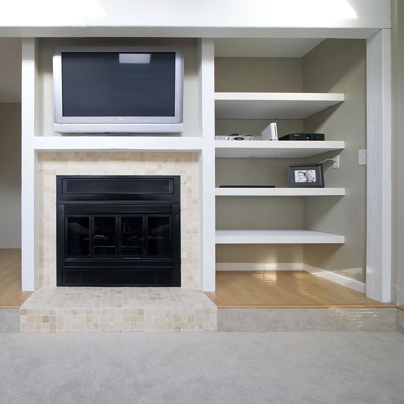 A fireplace with a black interior and tile hood, flanked by white floating shelves displaying framed photos, books, and decorative items.