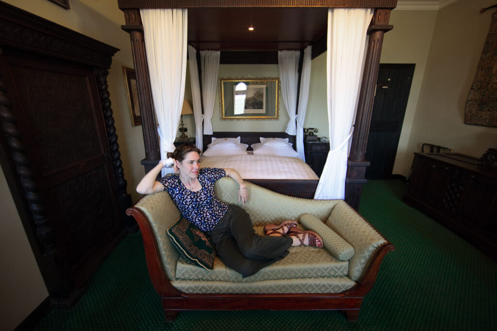 A woman lounges on a couch in a hotel room with four-poster beds and green carpeting. The space is warmly lit with classic decor.