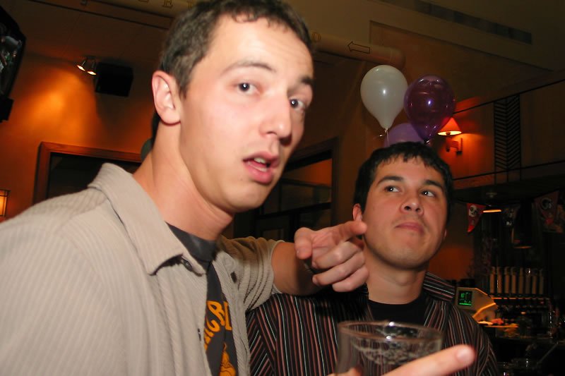 Two men are captured in a warm, dimly lit room, visibly enjoying each other's company. Balloons float in the background while one man points playfully at the other who holds a drink.