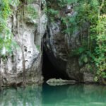 San Ignacio A crocodile rests near a cave entrance surrounded by lush greenery and calm water.