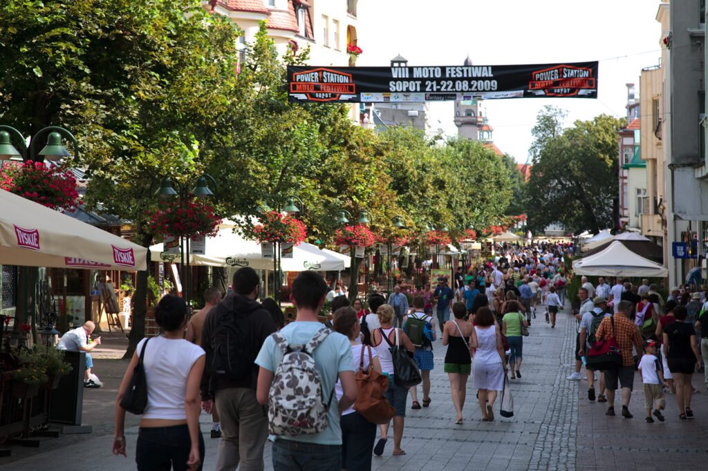 A lively street scene with crowds of people walking under a festival banner. The area is lined with trees and outdoor seating.