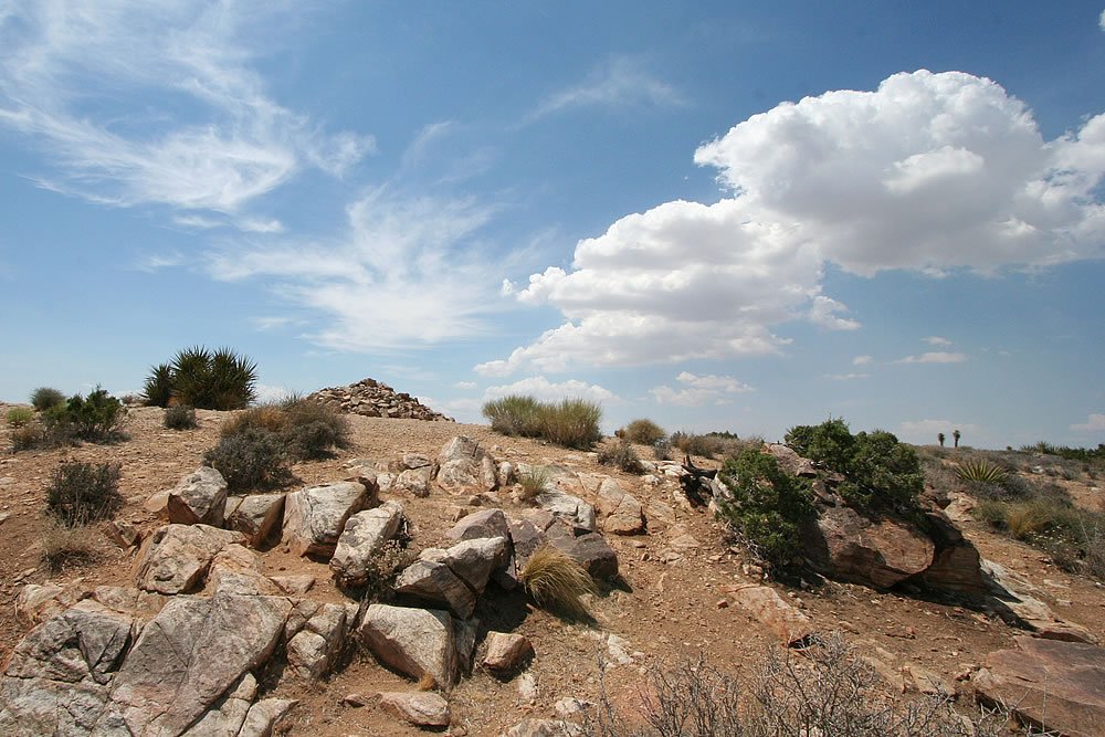 A rugged desert landscape under a wide sky, dotted with clouds and sparse vegetation.