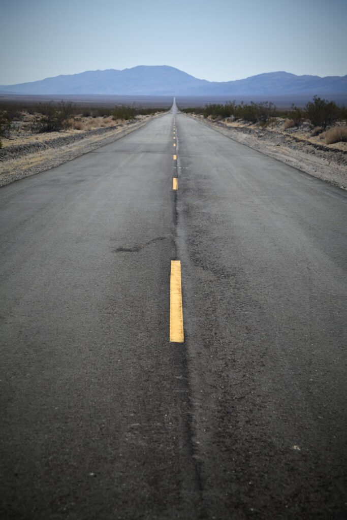A paved road with yellow center lines leading into a hazy desert landscape with distant blue mountains under a pale sky.