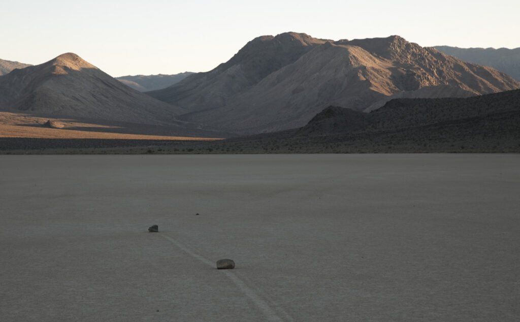 Two cars driving along a desert landscape, leaving tire tracks on the dusty surface, with large mountains in the background.