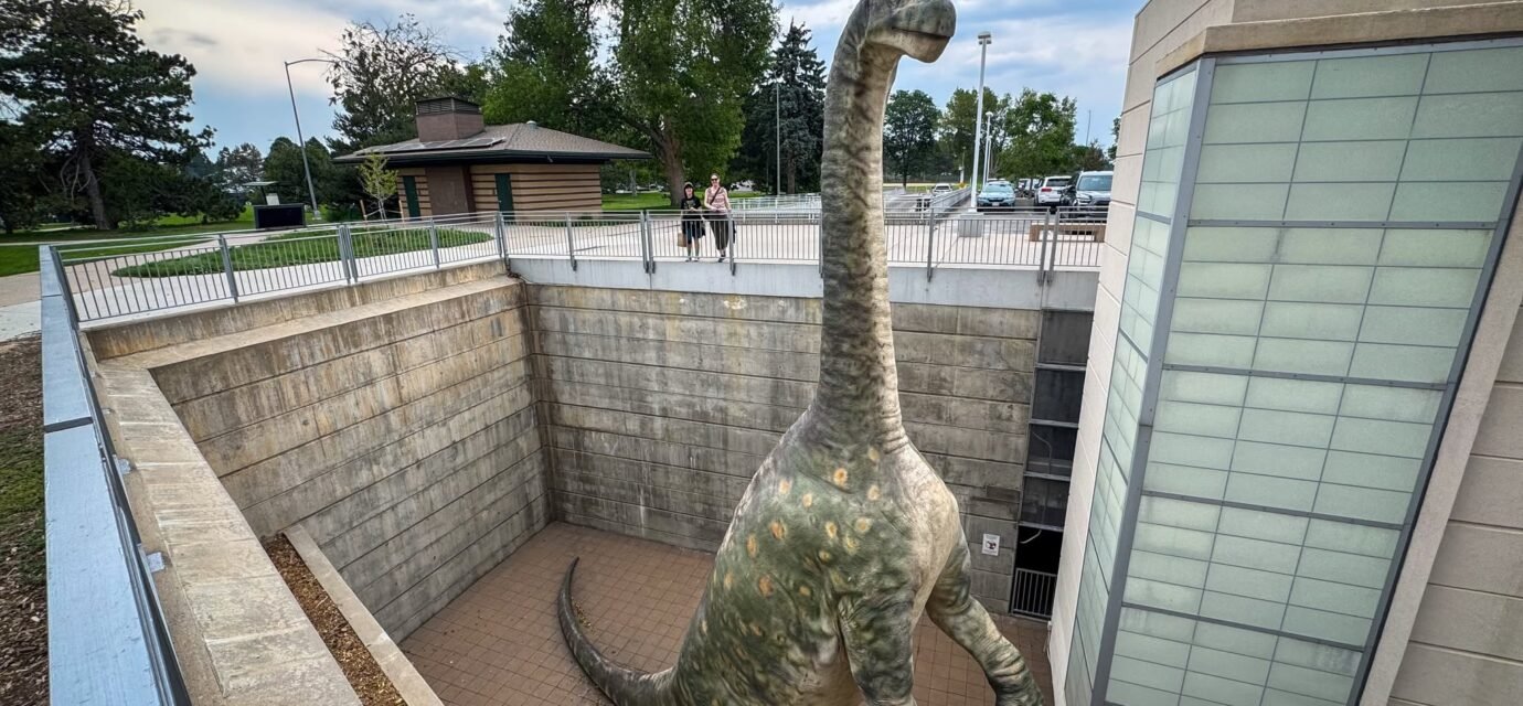 A large dinosaur illustration stands prominently inside a building, surrounded by windows and modern architectural elements.