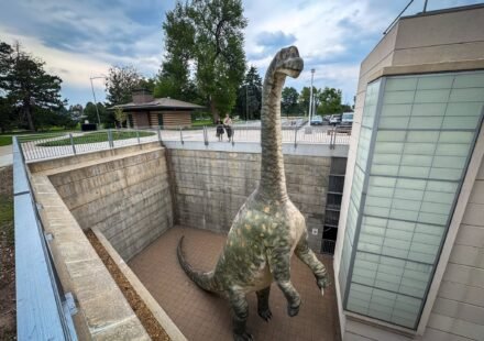 Denver Museum of Nature & Science A large dinosaur illustration stands prominently inside a building, surrounded by windows and modern architectural elements.