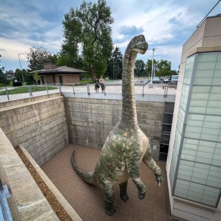 A large dinosaur illustration stands prominently inside a building, surrounded by windows and modern architectural elements.
