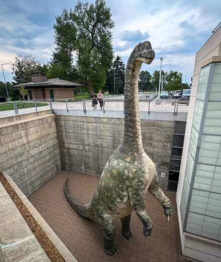 A large dinosaur illustration stands prominently inside a building, surrounded by windows and modern architectural elements.