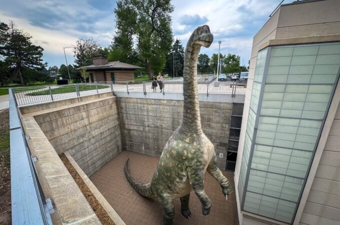A large dinosaur illustration stands prominently inside a building, surrounded by windows and modern architectural elements.