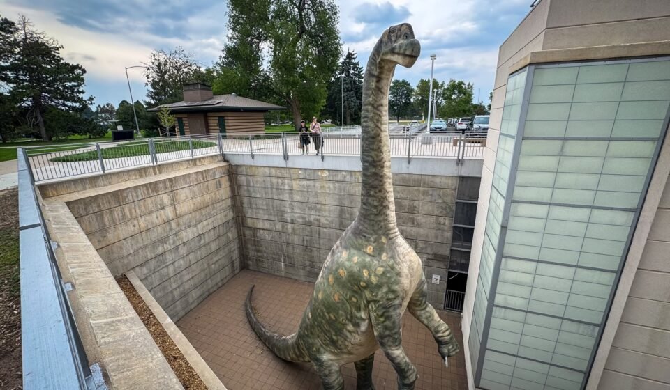A large dinosaur illustration stands prominently inside a building, surrounded by windows and modern architectural elements.