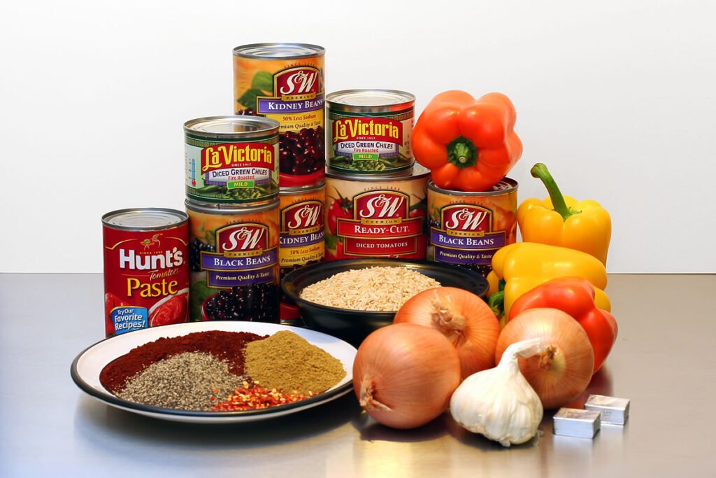 A colorful assortment of cooking ingredients including tinned goods, bell peppers, onions, garlic, and various spices displayed on a table.