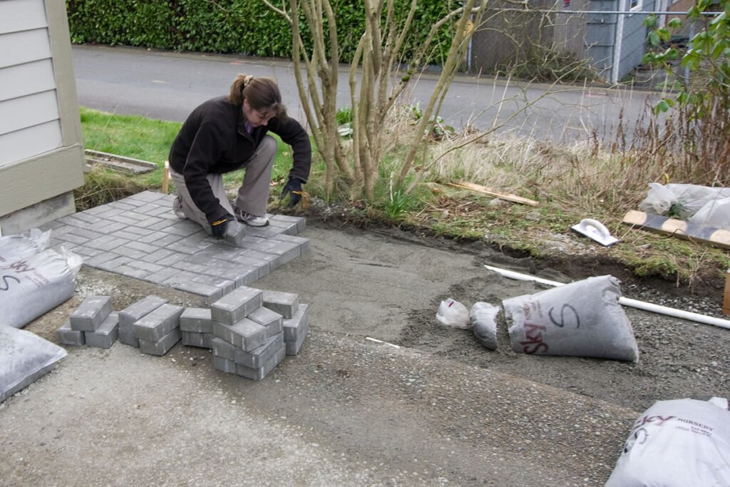 A person lays paving stones next to bags of gravel. Work tools are visible on the ground.