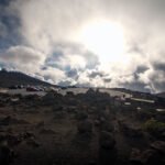 Haleakala A panoramic view of a dramatic sky with scattered clouds above a rugged landscape of rocks and dirt. The sun shines brightly through the clouds.