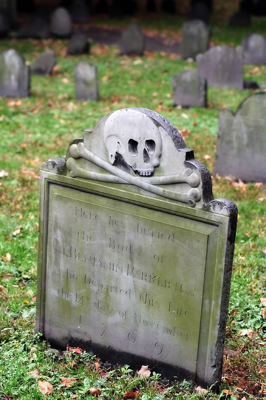 A historic tombstone topped with a carved skull and crossbones stands in a lush graveyard surrounded by autumn leaves.