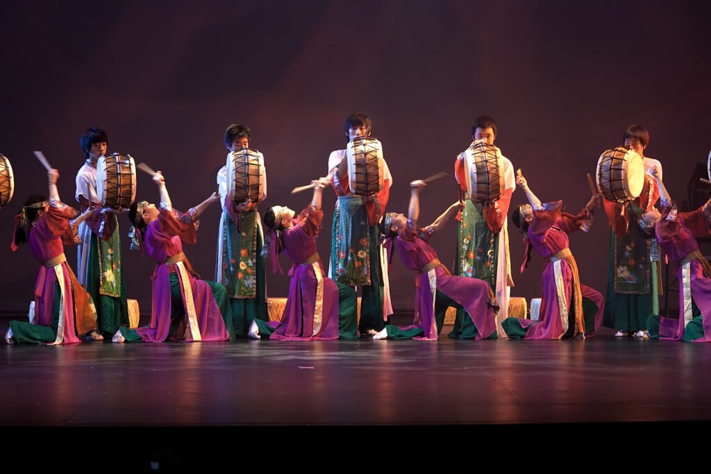 A group of performers in colorful costumes playing drums on stage with green and purple outfits, showcasing energetic movements.