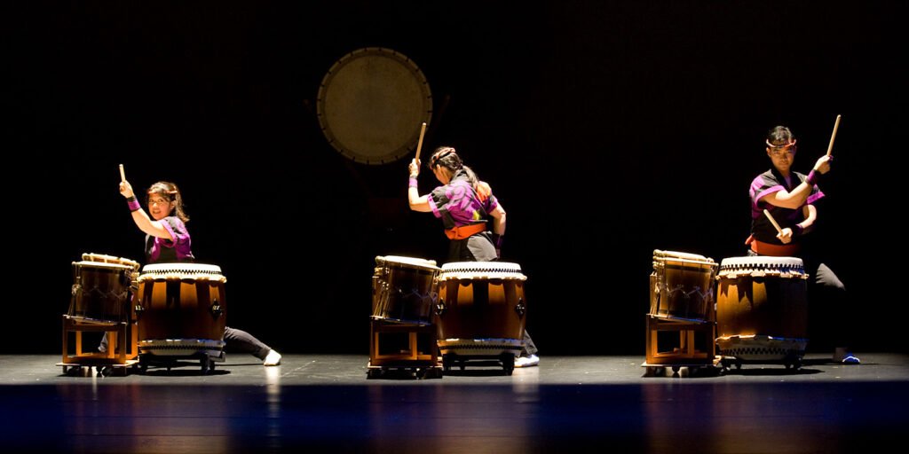 A group of drummers playing on stage with large gongs in the background.