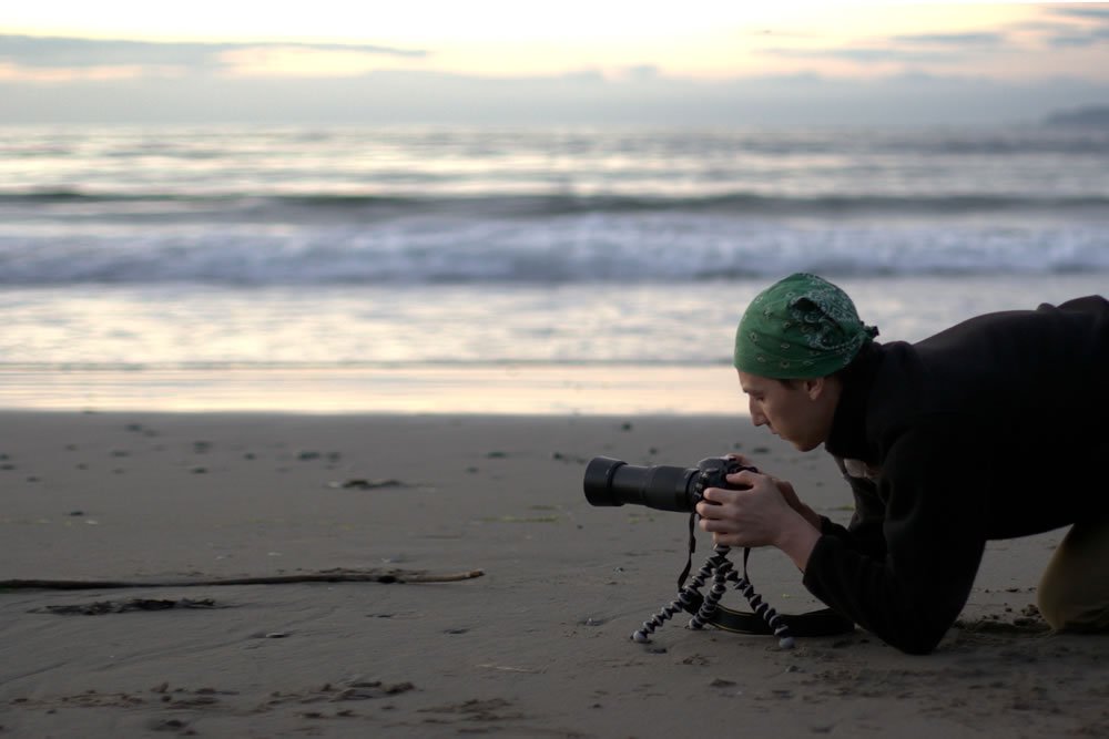 A photographer lies on the sand, capturing the ocean's beauty during dusk.