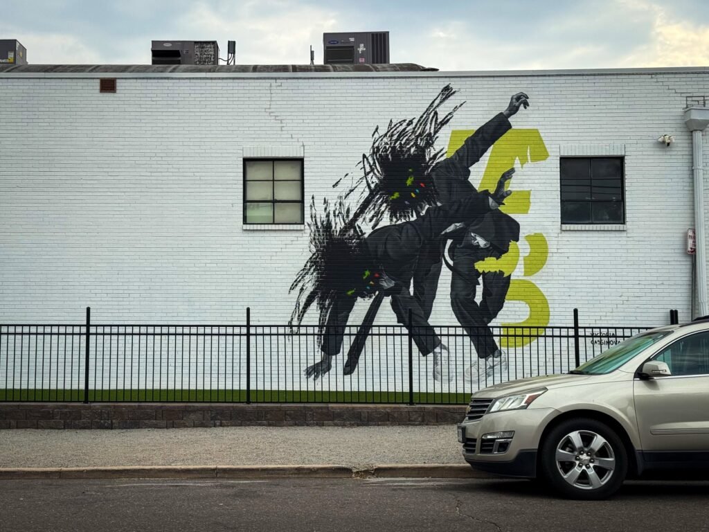 A striking mural of a leaping figure painted on a white building wall, with a parked car in the foreground.