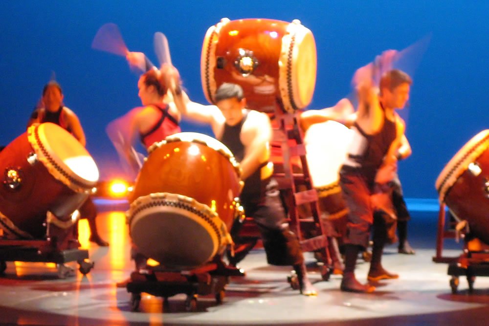 A group of performers energetically playing large drums on stage with a blue backdrop.