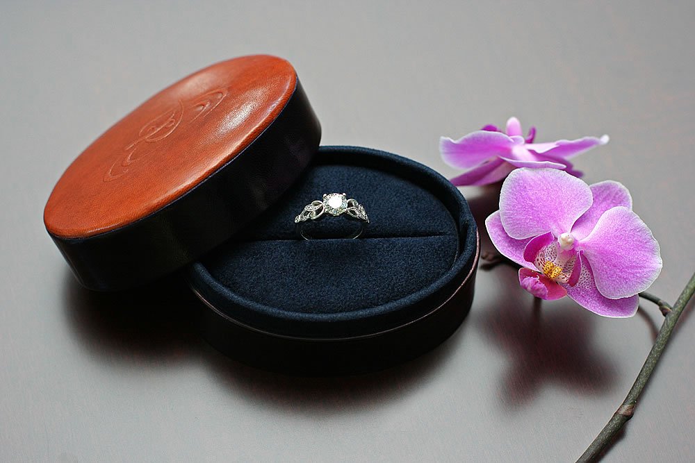 An elegant engagement ring rests in an open dark blue box, accentuated by a delicate pink orchid nearby.