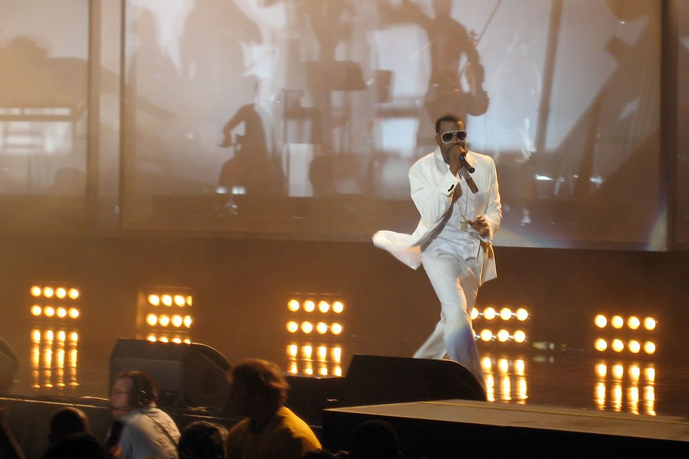 Elvis Presley in a white jumpsuit performing on stage with bright stage lights in the background. The audience is visible in the foreground, watching the performance.