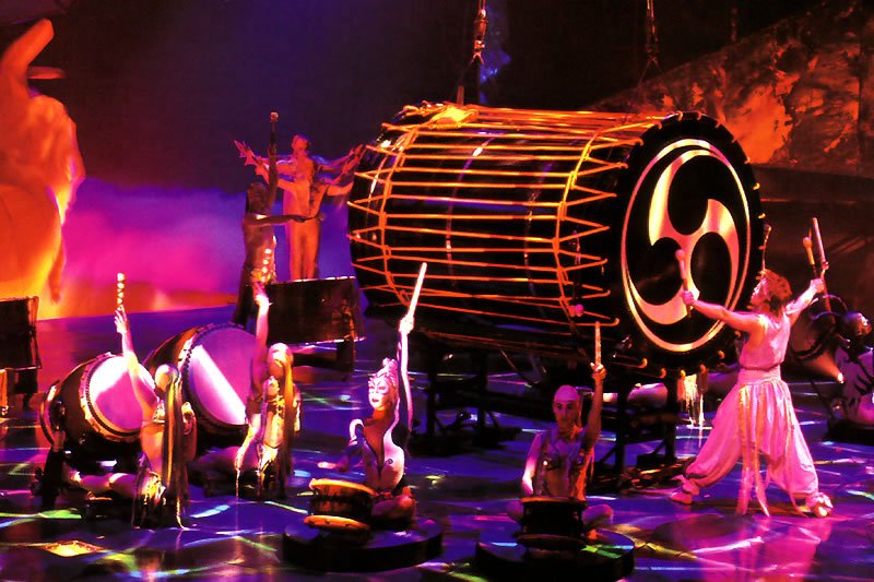 A vibrant stage performance featuring musicians playing large drums under colorful lighting.