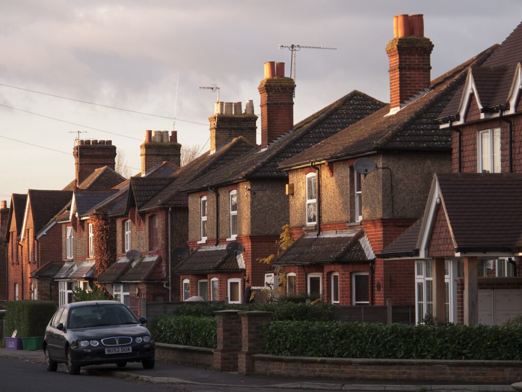 A scenic view of a suburban street lined with red brick houses, featuring a parked car and neatly trimmed hedges.