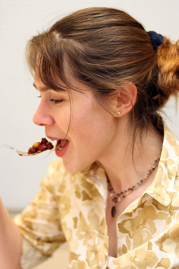 A woman in a patterned blouse happily eating a forkful of food. She has her hair tied back and wears a simple necklace.