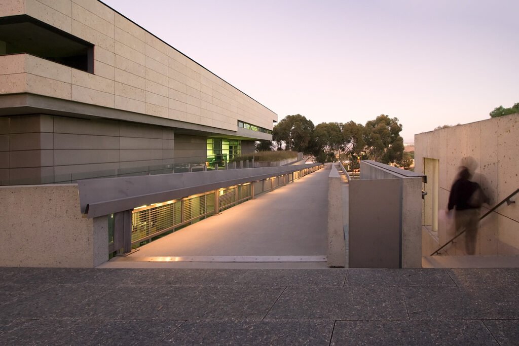 A person walks up a modern concrete ramp under the soft glow of evening sky, framed by clean architectural lines and distant trees.