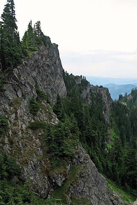 The edge of a mountain ridge with rugged rock formations and dense evergreen trees. In the distance, layers of blue hills fade into the horizon under a cloudy sky.